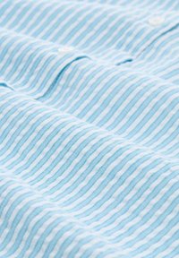 Tessuto a righe blu chiaro con sottili linee bianche alternate. Presenta una texture morbida e piccoli bottoni bianchi visibili nelle pieghe.