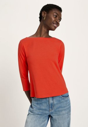 Woman with short curly hair wearing gold hoop earrings, a bright red long-sleeve top, and light blue jeans, smiling and looking right.