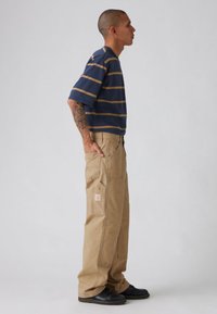 Μπλούζα με κοντά μανίκια σε ριγέ ναυτικό μπλε και καφέ, συνδυασμένη με μπεζ παντελόνι cargo με πολλές τσέπες και ετικέτα, φορεμένη με μαύρα παπούτσια.