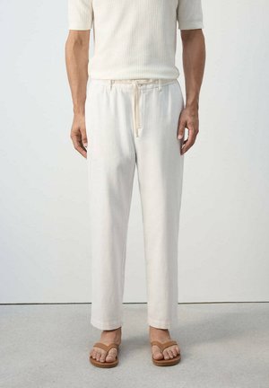 Hombre con pantalones color crema con cordón, camiseta de manga corta texturizada y sandalias marrones, de pie frente a un fondo liso.