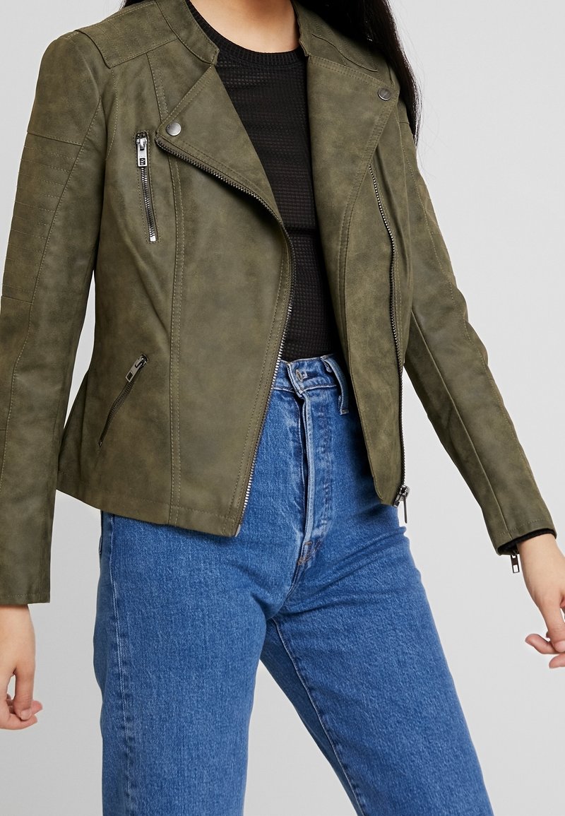 Veste de moto en simili cuir vert olive avec épaulettes, fermeture éclair asymétrique et deux poches avant zippées, associée à un jean taille haute.