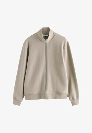 Beige ribstrikket jakke med lynlås, lange ærmer og en høj krave, med en synlig sølvlynlås foran.