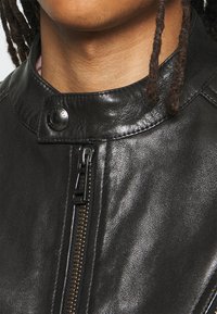 Black leather jacket featuring a stand-up collar, zip closure, and snap button detail. Texture is smooth with slight sheen.