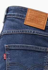 Gros plan sur un jean en denim bleu Levi's avec poche arrière, coutures visibles, étiquette rouge Levi's et étiquette de marque en cuir marron sur la ceinture.