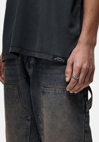 T-shirt grigio scuro abbinato a pantaloni di denim blu sbiadito. I pantaloni presentano tasche laterali e un'etichetta di marca visibile, con un anello metallico indossato sulla mano.
