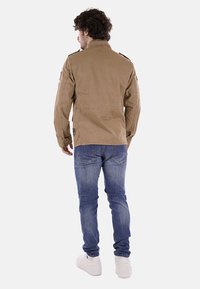 Giacca color khaki con chiusura a zip, spalline sulle spalle e tasche laterali, abbinata a jeans blu e sneakers bianche. Tessuto testurizzato.