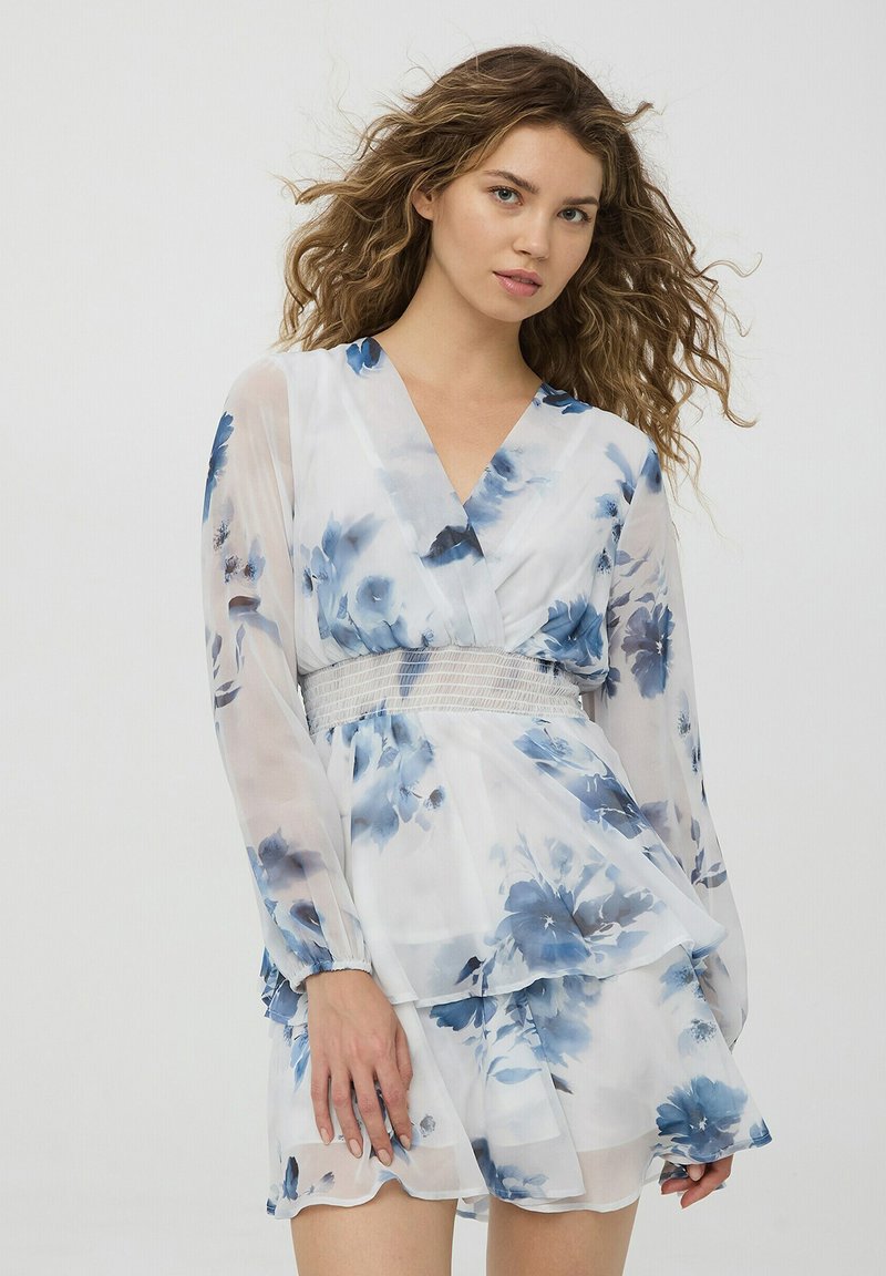 Abito floreale in tessuto bianco trasparente con fiori blu. Presenta maniche lunghe, scollatura a V profonda e vita smock. Design a gonna sovrapposta.