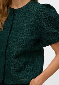 Tume roheline tekstuuriga pluus ümarate kaeluste ja lühikeste puffvarrukatega. Omab struktureeritud kortsulist materjali ja ees olevat nööpide sulgumist.