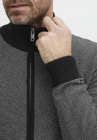 Sudadera con cremallera en negro y gris, con un alto cuello, que presenta un patrón de rayas texturadas y puños de canalé, decorada con una cremallera plateada.
