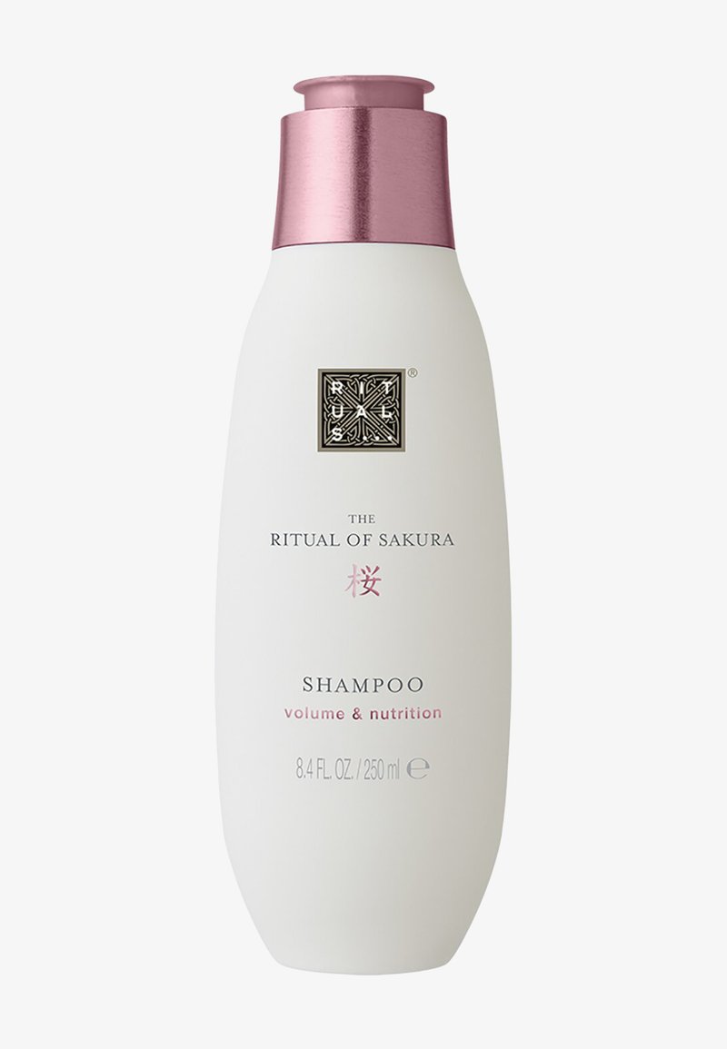 Botella blanca con forma curvada, tapa metálica rosa y logotipo en relieve. La etiqueta indica "El Ritual del Sakura" champú para volumen y nutrición.