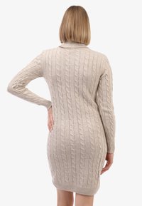 Beige kudumekleid, millel on nöörmustrit, pikad varrukad, kõrge krae ja figuuri järgiv siluett, mis lõpeb puusade keskel. Tekstuuriga kangast detail on nähtav.