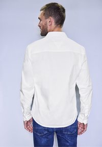 Camicia bianca a maniche lunghe con colletto, texture liscia e polsini abbottonati, abbinata a jeans blu, vista da dietro.