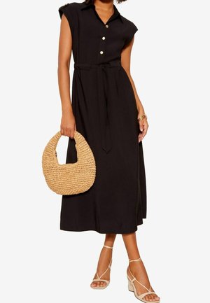 Schwarzes Kleid mit kurzen Ärmeln, Knöpfen vorne, einer Taillenbindung und mittlerer Länge. Kombiniert mit einer gewebten, hellbraunen Handtasche und zarten, nude-farbenen Sandaletten.