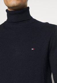Maglione a collo alto blu navy con un piccolo logo rettangolare rosso, bianco e blu sul lato sinistro del petto, indossato da una persona.