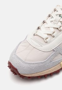 Beige sneakers med grå semsket skinn aksenter, teksturert gummisåle i rød, forsterket tåhette, og snøring med polstret tunge.