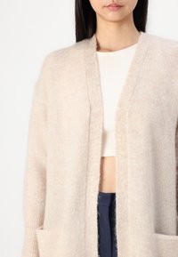 Světlý béžový kardigan s měkkou, chlupatou strukturou. Má šálový límec, otevřený předek a dvě přední kapsy. Nošený přes bílé tričko.