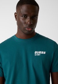 Teal cotton t-shirt featuring a crew neckline and short sleeves. White "GUESS" logo with "©1981" on left chest. Smooth texture.
