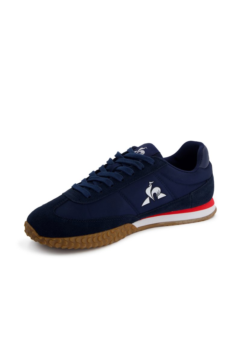 Zapatilla deportiva azul oscuro con parte superior de ante y sintético, suela de goma, media suela a rayas blanca y roja, que presenta un logotipo en el costado.