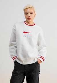 Sivi dukserica s crvenim detaljima i okruglim izrezom. Ima „NIKE“ logo u podebljanoj crvenoj boji i godište „1972“ izvezeno na prednjem dijelu.