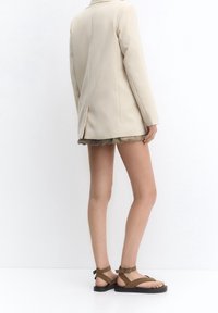 Beige op maat gemaakte blazer met een enkele split, gecombineerd met olijfgroene shorts met ruches en bruine platte sandalen met enkelbandjes.