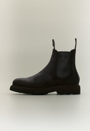Botte Chelsea en cuir noir avec panneau latéral élastique, languettes à l'avant et à l'arrière, et semelle texturée sur fond clair.