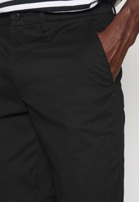 Pantalon en coton noir avec une texture lisse. Caractérisé par une fermeture à bouton et des poches latérales, mettant en valeur une coupe ajustée.