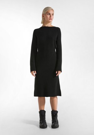 Abito nero lavorato a maglia con scollatura a barca, maniche lunghe e silhouette aderente, abbinato a robusti stivali neri con lacci.