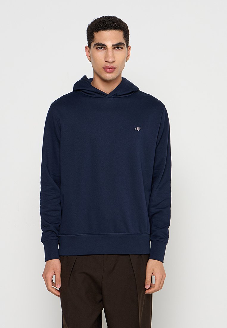 Marineblaue Kapuzenjacke aus weichem Stoff, mit einer vorderen Kängurutasche und gerippten Bündchen. Dezentes Branding auf der Brust.