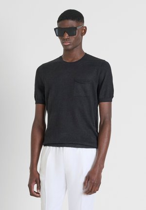 T-shirt nera a maniche corte in maglia leggera, con una piccola tasca sul petto, abbinata a pantaloni bianchi e occhiali da sole oversize neri.
