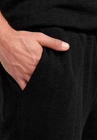 Schwarze, strukturierte Loungewear-Hosen mit einer Hand in der Tasche. Der Stoff hat ein Waffelmuster und verfügt über einen elastischen Bund für zusätzlichen Komfort.