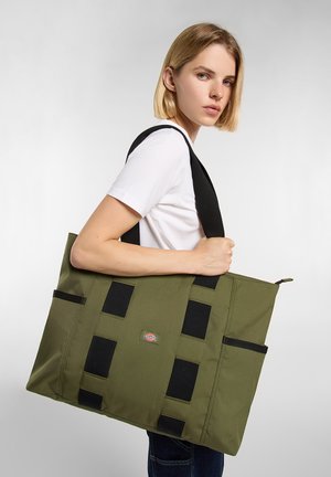 Torba materiałowa w kolorze oliwkowej zieleni z czarnymi paskami, wyposażona w cztery czarne akcenty i zamknięcie na suwak. Na przodzie widoczne charakterystyczne logo marki.