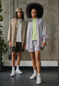 Två modeller står framför en betongvägg. Den ena bär en beige oversize-kappa, shorts och vita sneakers. Den andra bär en randig skjorta och shorts.