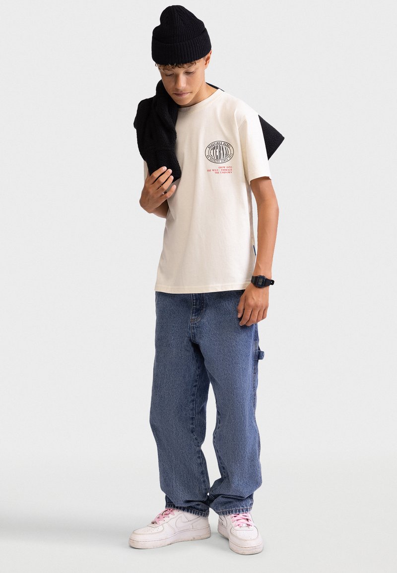 Beige bomulds T-shirt med rundt logo, blå denim bukser, sort strikket beanie, sort sweater over skulderen, hvide sneakers med lyserøde snørebånd.