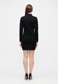 Γυναίκα που στέκεται με την πλάτη προς τα εμπρός, φοράει στενό μαύρο μικρό φόρεμα με μακριά μανίκια και διακριτικό μοτίβο, καθώς και μαύρα ψηλοτάκουνα παπούτσια σε λευκό φόντο.