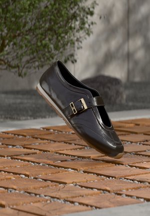 Mocassin en cuir marron foncé avec boucle dorée flottant au-dessus d'un sol en bois et pierre à motifs, avec un buisson vert flou en arrière-plan.