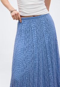 Γυναίκα που ρυθμίζει τη μέση μιας υφασμάτινης μπλε μίντι φούστας, φορώντας λευκό στρωμένο τοπ και ασημένιο βραχιόλι με κόμπους στον αριστερό καρπό.