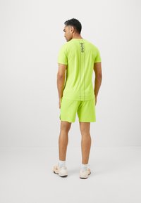 Hombre vistiendo ropa deportiva de color verde lima con el texto "BORG" en la parte posterior, de pie y mirando hacia un lado frente a un fondo blanco liso.
