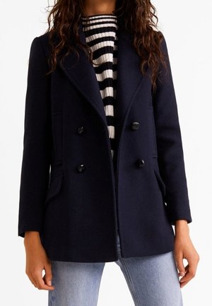 Vrouw draagt een zwarte dubbelgeknopte jas over een zwart-wit gestreepte trui en lichtblauwe spijkerbroek.