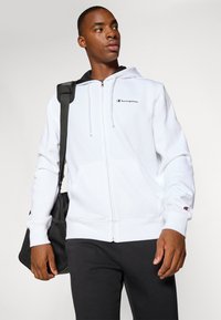 Witte ritssluitingen hoodie van een katoenmix, met een zwart logo op de borst, trekkoorden en zijzakken. Model draagt een zwarte tas.