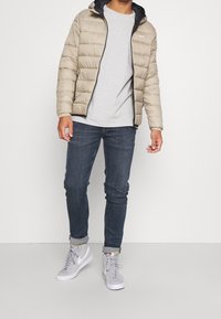 Beżowa puchowa kurtka z kapturem i czarną podszewką, szara koszulka z okrągłym dekoltem, ciemnoniebieskie dżinsy oraz szare sneakersy z białymi akcentami.