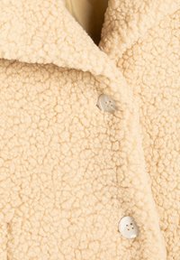 Abrigo de forro polar beige con una superficie texturizada, gran cuello con muesca y botones blancos para el cierre. Material suave y cálido con un tacto de felpa.