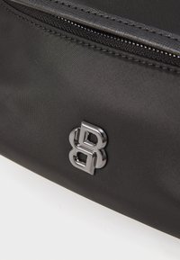 Crna tkanina torba s teksturiranom površinom, koja ima zatvaranje na zip i metalni logo kao naglasak. Dizajn je minimalistički i suvremen.