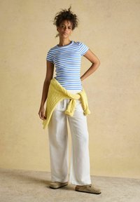 Kobieta stojąca z ręką na biodrze, ubrana w niebiesko-białą koszulę w paski, białe spodnie o szerokich nogawkach, beżowe buty wsuwane oraz żółty sweter przewiązany wokół talii.