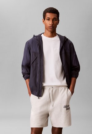 Homme portant une veste à capuche foncée sur un t-shirt blanc et un short blanc Calvin Klein, debout les mains dans les poches sur un fond uni.