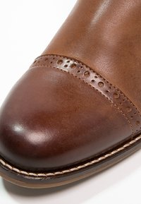 Brązowy skórzany but o zaokrąglonym nosku, z perforowanym akcentem i kontrastującym ciemnobrązowym nosku. Widoczne detale szycia.