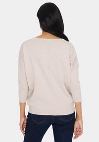 Hellbeige, oversized Strickpullover mit Fledermausärmel, überschnittenen Schultern, geripptem Saum und mittigem Nahtdetail, kombiniert mit dunklen Jeans.