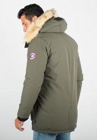 Parka vert olive avec capuche en fourrure, poches latérales et poignets côtelés. Présente un patch logo circulaire sur le bras gauche.
