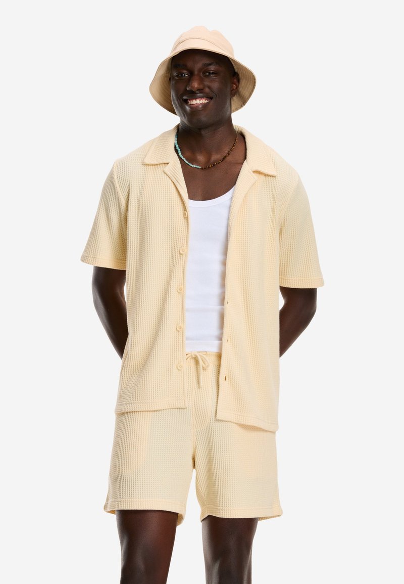 Smilende mand iført en lys beige kortærmet skjorte med knapper, matchende shorts, hvid tanktop, beige bucket hat og en perlekæde.
