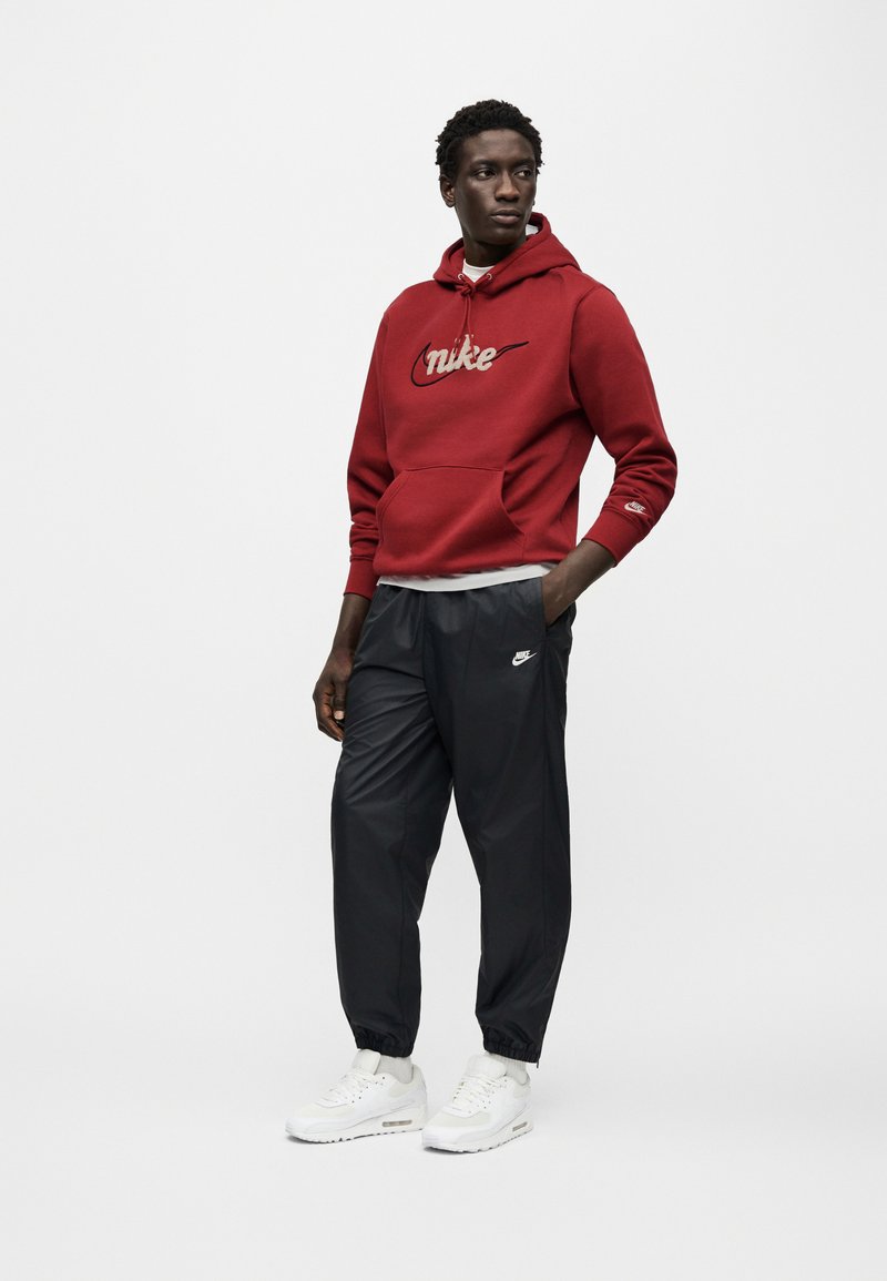 Felpa rossa con cappuccio con logo Nike beige, tasca frontale e polsini a coste. Pantaloni sportivi neri e scarpe da ginnastica bianche con ammortizzazione visibile ad aria.