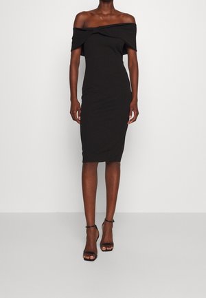 Femme portant une robe noire ajustée épaules dénudées longueur genoux et des sandales noires à talons hauts avec bride à la cheville, debout sur un fond blanc.